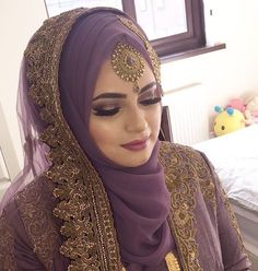 a woman wearing a purple hijab and gold jewelry looks down at her cell phone