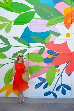 a woman in a red dress and hat standing next to a colorful wall with flowers on it