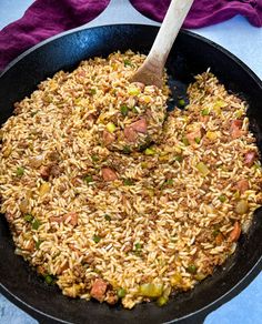 a skillet filled with rice and vegetables on top of a blue table cloth next to a wooden spoon