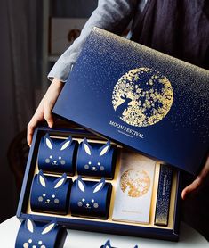 a person holding a blue box with gold foiled designs on it and six pieces of chocolate