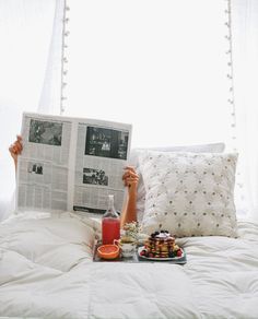 a person laying in bed reading a newspaper and holding up a plate with food on it