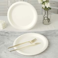 two white plates with gold forks are sitting on a gray table cloth next to a vase