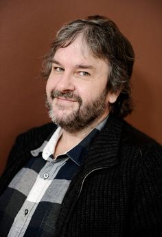 a man with long hair and beard wearing a black jacket smiles at the camera while standing in front of a brown wall