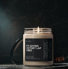 a candle sitting on top of a table next to a black mug with the words it's giving victory lap vibes