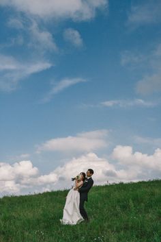 a bride and groom standing in the grass under a blue sky with white fluffy clouds