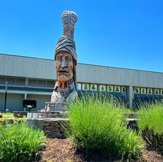 a large wooden statue in front of a building with green plants and bushes around it