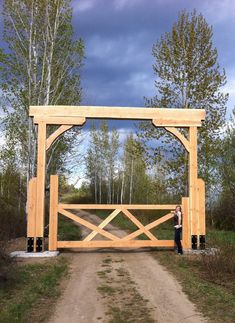 a man standing in front of a wooden gate on a dirt road with trees behind him