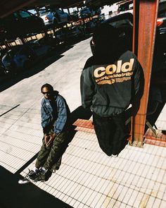 two young men standing next to each other on a sidewalk with cars parked in the background