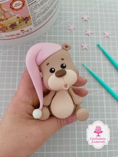 a hand holding a small teddy bear with a pink hat on it's head