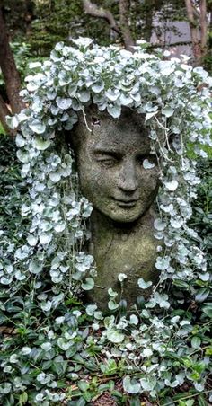 a statue in the middle of some bushes and plants with flowers growing on it's head