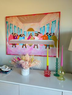 a painting on the wall above a table with candles and flowers in front of it