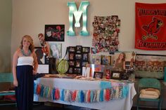 a woman standing in front of a table with many items on it and the letter m above her