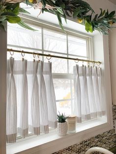 a kitchen window with white curtains and green plants hanging from the windowsill, next to a potted plant