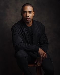 a man sitting on top of a wooden stool in front of a dark background with his arms crossed