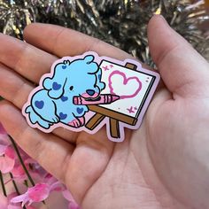 a hand holding a blue sticker with a heart on it and an easel in the background