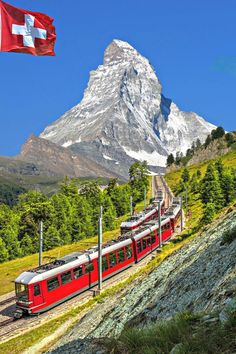 a red train traveling past a mountain with a flag on it's back end