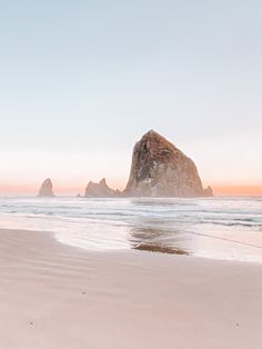 two people walking on the beach with an ocean and rock in the background at sunset