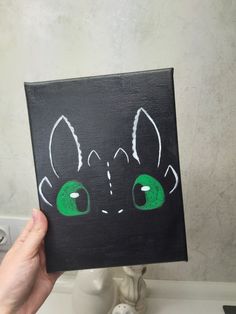 a hand holding up a black and green painting with an image of a cat's face on it