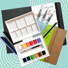 an assortment of art supplies including watercolors, pens and paper on a blue background