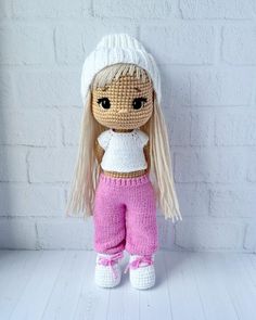 a crocheted doll with blonde hair wearing pink pants and a white top is standing against a brick wall