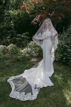 a woman in a wedding dress and veil standing on the grass with her back to the camera