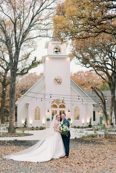 a bride and groom standing in front of a church with fall leaves on the ground