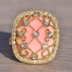 The 154th Avenue: Pink Coral And Natural Diamond Cocktail Ring In Yellow Gold. The Ring's Focal Point Is A Central Round-Cut Natural Diamond Ensconced Within A Lattice Of Yellow Gold Lace Adorned With Natural Diamonds. Below This Intricate Design Rests A Cushion Buff Top Made Of Exquisite Pink Coral. Encircling The Coral Is A Halo Of Round-Cut Natural Diamonds. The Split Shank Of The Ring Is Also Adorned With Round Brilliant-Cut Natural Diamonds. Delicate Milgrain Detailing Graces The Lacework, Both Outside The Halo And Around The Bezels. Crafted In 14 Karat Yellow Gold, The Ring Is Currently Sized At 6.75 But Can Be Resized To Fit Any Finger Size For An Additional Charge Upon Request. Luxury Pink Diamond Ring For Formal Occasions, Luxury Pink Oval Diamond Ring, Luxury Pink Diamond Gemstone Ring, Pink Diamond Ring With Pave Setting For Gift, Pink Diamond Ring With Pave Setting As Gift, Luxury Pink Diamond Ring With Single Cut Diamonds, Exquisite Pink Diamond Ring With Accents, Elegant Pink Diamond Ring With Diamond Accents, Luxury Pink Jewelry With Prong Setting