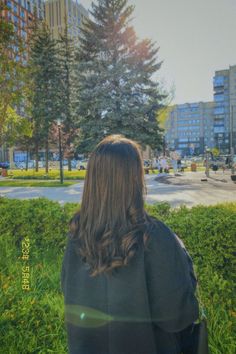 a woman is sitting in the grass looking at some buildings and trees on either side of her