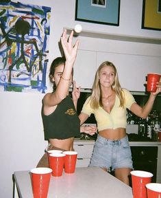 two young women standing in a kitchen holding cups and throwing a frisbee into the air