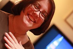 a woman wearing glasses and smiling in front of a computer monitor with her hands on her chest