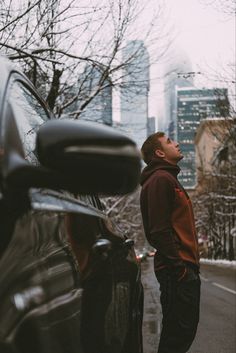 a man standing next to a car looking up at the sky with buildings in the background