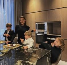 three children sitting at a kitchen table with food in front of them and an adult standing behind them