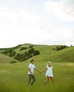 a man and woman holding hands while walking through the grass in an open field with rolling hills behind them