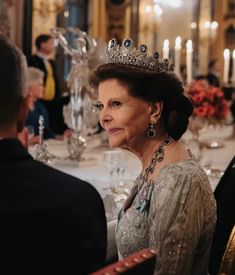 an older woman wearing a tiara sitting at a table with other people in formal wear