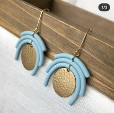 two tone blue and gold earrings hanging from a wooden hook on a white fabric background