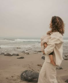 a woman holding a baby on the beach with rocks in the foreground and water in the background