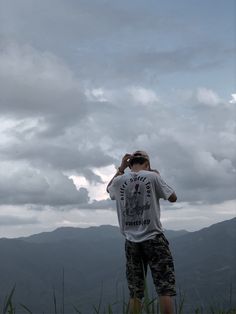 a man standing on top of a lush green hillside under a cloudy sky with mountains in the background