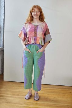 a woman standing in front of a white wall wearing green pants and a colorful top