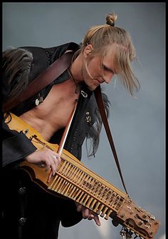 a man with long hair holding a wooden instrument on top of his chest and looking down at the ground