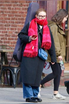 a woman walking down the street with a guitar case in her hand and a red scarf around her neck