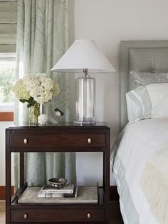 a nightstand with flowers on it next to a lamp and bed in a room that has green curtains