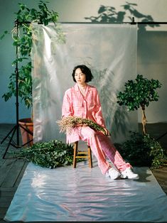 a woman sitting on top of a chair in front of a white backdrop and potted plants