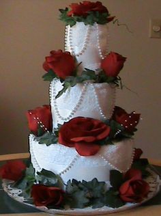 a three tiered white wedding cake with red roses on the side and pearls around the edges