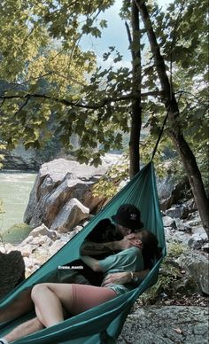 two people laying in a hammock on the river bank with their backs to each other