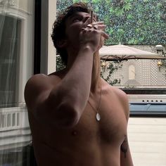 a shirtless man standing in front of a window talking on a cell phone while holding his hand up to his face