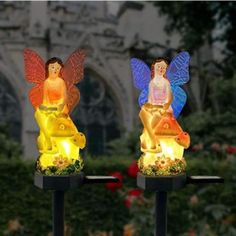 two colorful fairy lights sitting on top of black stands in front of bushes and flowers