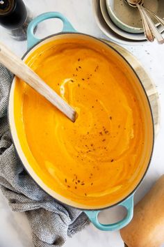 a blue pot filled with carrot soup next to two butternuts and a wooden spoon