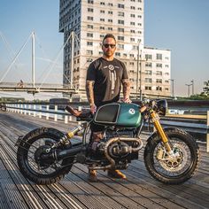 a man standing next to a motorcycle on a wooden platform with buildings in the background