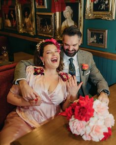 a man and woman sitting at a table with flowers in their hair, laughing for the camera