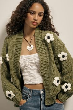 a woman wearing a green cardigan with white flowers on the sleeves and back, standing in front of a wall